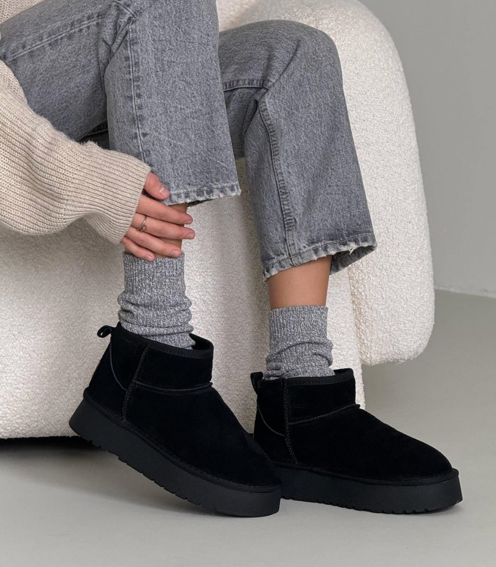 Black suede ankle boots paired with gray jeans and textured gray socks on a cozy chair.